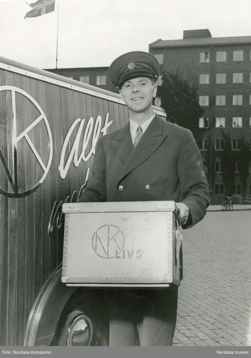 En leende, uniformsklädd man med Nordiska Kompaniets varumärke i skärmmössan håller i en trälåda med texten "NK-LIVS". Bredvid honom en NK-märkt bil. Text med blyerts på baksidan: "Nya uniformer NK:s varubilar."