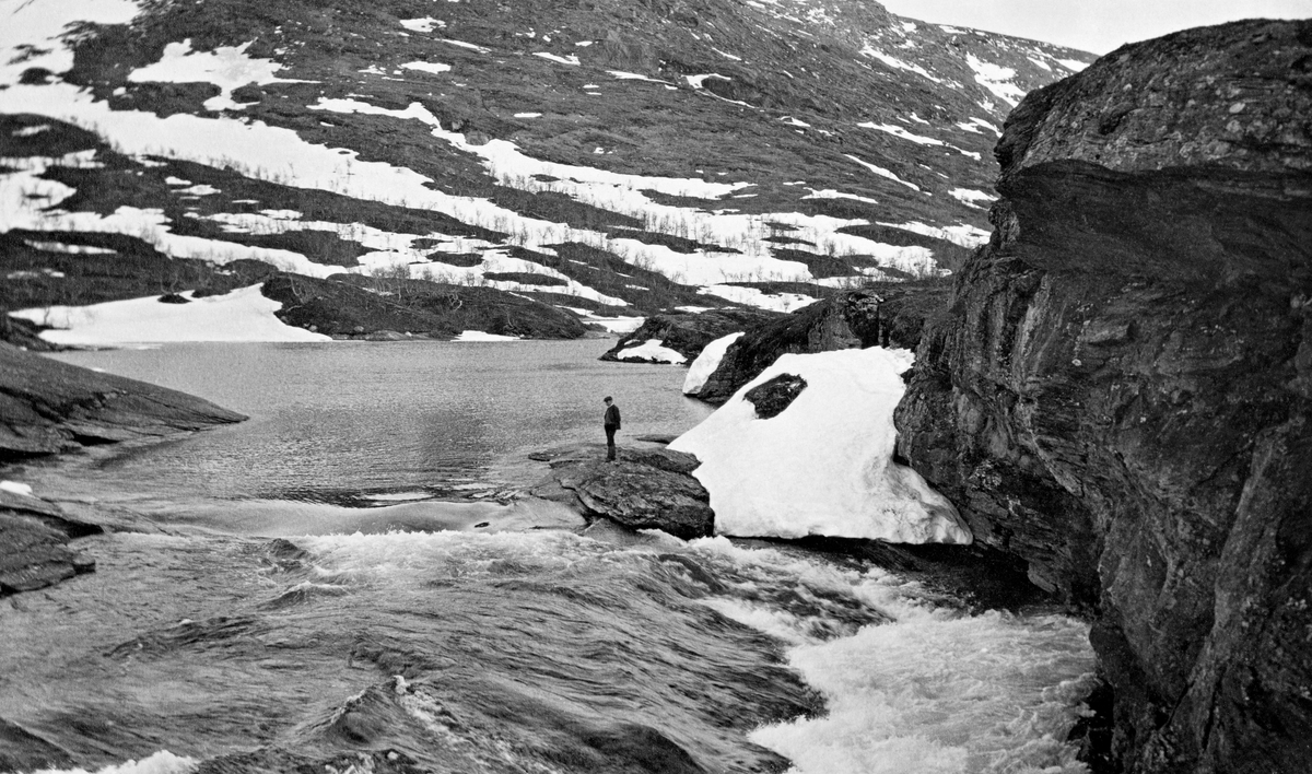 Utløpet av Kjennsvatnet i Bjerkavassdraget i Hemnes kommune i Nordland. På fotografiet ser en hvordan elva renner ut av vatnet mot en bergsprekk ved en bratt bergrygg.  En mann står på et steinskjær ved innløpet til fossen.  Dette stedet har tydeligvis fotografen fra Vassdragsvesenet sett som et potensielt damsted.  I bakgrunnen ei fjellside med snøstrimer og småbjørk.