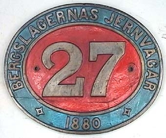 Oval skylt av mässing med text i relief mot röd- och blåmålad botten.
Från ångloket BJ K 27. Loket såldes till en skrothandlare 1947 som i sin tur sålde loket till SJ 1948, 1956 kom loket till Sveriges Järnvägsmuseum.
Beyer Peacock & Co. Nº 1933.