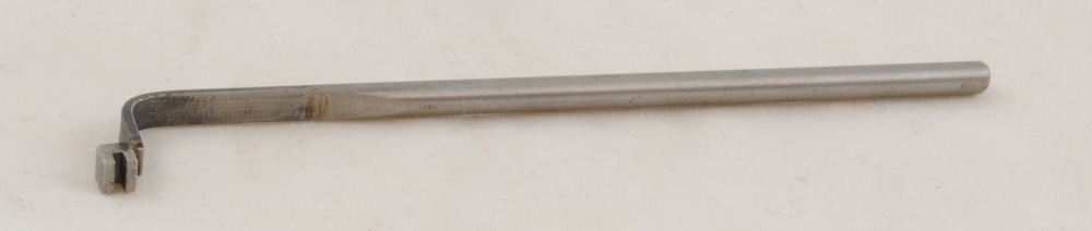 Verktyg (fjäderbockare?) bestående av en metallpinne som är krökt och platt i ena änden med en rektangulär metallbit med en skåra.