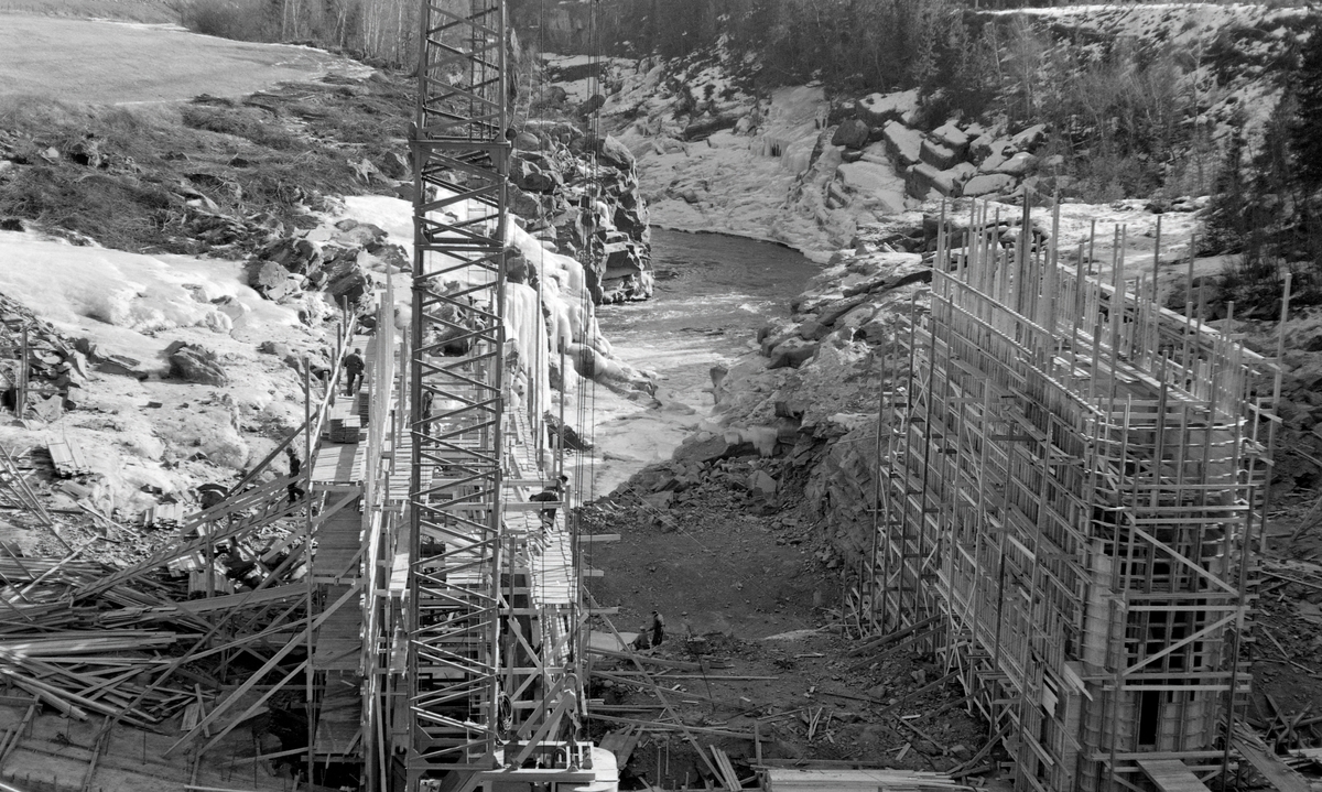 Anleggsarbeid i forbindelse med kraftutbygginga i Harpefossen i Gudbrandsdalslågen - i Sør-Fron kommune i Oppland fylke - vinteren 1964.  Fotografiet er tatt fra brua over elva ved Solbrå, like ovenfor fossen.  Vi ser ned på et anleggsområde der man var i ferd med å reise forskalinger for to digre betongkar for kraftverksdammen.  I bakgrunnen ser vi noe av det trange elvegjelet der elvevatnet i flomtider ble slynget høyt oppover de bratte sideveggene.  Den kraftige strømmen og løsmasser som fulgte med vannet førte til at det dannet seg jettegryter, glatte, runde forsenkninger i berget.  Harpefossen hadde vært et vanskelig punkt for tømmerfløterne, og når tømmeret satte seg fast der, var det meget farefullt arbeid å løsne stokkene igjen.  Da dette fotografiet ble tatt var elveleiet langt på veg tørrlagt.  Kraftutbygginga endret vannføringa i den nærmest nedenforliggende delen av Lågen radikalt.  Kraftverket utnytter et fall på 34 meter.  Kraftstasjonen ved Harpefossen har to kaplanturbiner og en årsproduksjon på 427 GWh.  Den ble satt i drift i 1965, året etter at dette fotografiet ble tatt.