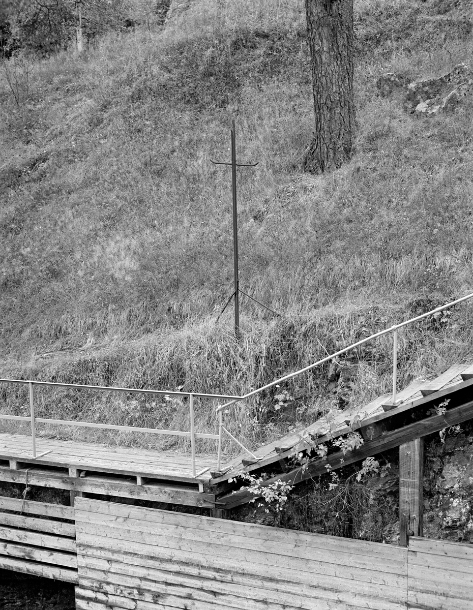 Detalj fra sluseanlegget ved Ørje i Marker kommune i Østfold.  Fotografiet er tatt i 1982, som var den siste fløtingssesongen i Haldenvassdraget.  Bildet er tatt på tvers av kanalløpet.  Vi ser ei tretrapp som førte fra gangbanen langs ovenforliggende slusekammer til gangbanen langs det nedenforliggende.  Så vel trappa som gangbanen hadde rekkverk av kvitmalte jernrør.  Hovedmotivet her må likevel ha vært jernstativet i den grasbevokste skråningen bakenfor.  Den skal ha tjent som oppheng for parafinlamper i tida før Ørje fikk elektrisk strøm til belysning.  Kanalen var i drift også på årstider da dagslysperioden var kort.

En liten historikk om tømmerfløting og kanaliseringsarbeid i Haldenvassdraget finnes under fanen «Opplysninger».