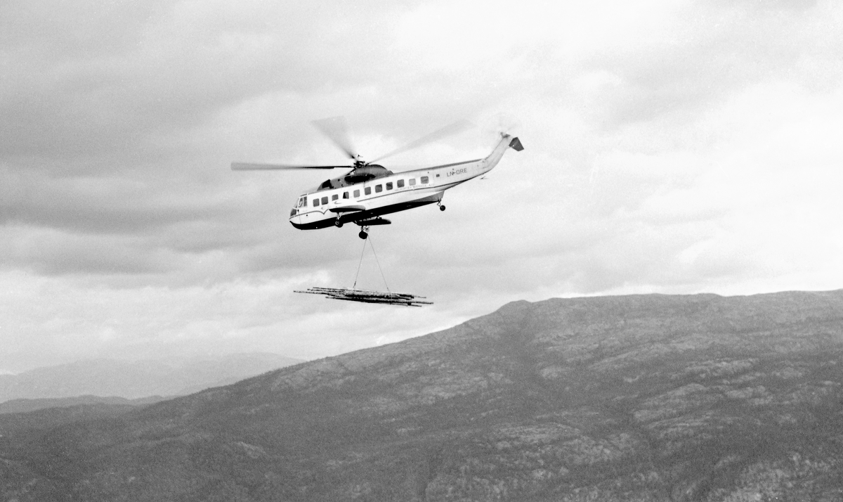 Helikopter av typen Sikorsky S 61 N, fotografert mot en skyet himmel med en bunt tømmer under transportforsøk nord for Dyrskarnut i Kviteseid i 1969.  Herfra skulle tømmeret farktes om lag 2,2 kilometer til en ringbom ved Mølen i Kviteseidvatnet, 650 høydemeter lavere i terrenget, hvor det kunne bukseres videre med slepebåt.  Dette amerikanskproduserte helikopteret hadde en motor som kunne yte 1 500 hestekrefter.  Maskinen var drøyt 18 meter lang (ekskl. hovedrotor) og veide drøyt 5,5 tonn.  Løftekapasiteten ble oppgitt å være 2,5 tonn, men forskerne vågde ikke å nytte denne kapasiteten fullt ut.  De nøyde seg av sikkerhetsgrunner med en utnyttingsgrad på om lag 70 prosent.  I praksis betydde dette at et gjennomsnittlig lass eller «hiv» ble på 2,95 kubikkmeter (fastmasse).  Det ble hentet 34 lass – om lag 100 kubikkmeter fastmasse – fra hogstfeltet ved Dyrskarnut ned til Kviteseidvatnet.  Arbeidet gikk raskere enn det hadde gjort i forsøkene med det langt mindre Bell-helikopteret man eksperimenterte med i Kviteseid tidlig i 1960-åra.  Timeprisen for bruk av Sikorsky-maskinen var imidlertid 7 150 kroner, og bortimot 30 prosent av tida gikk med til drivstoffylling og ettersyn av maskinen.  Konklusjonen ble derfor at «.. Slike timepriser fører til at helikoptertransport av tømmer blir langt kostbarere enn hvilken som helst annen tradisjonell tømmertransportmetode i fjellterreng.»  Forskerne mente at radiostyrte taubanedrifter, som de hadde investert mye utviklingsarbeid i, ville vært å foretrekke.