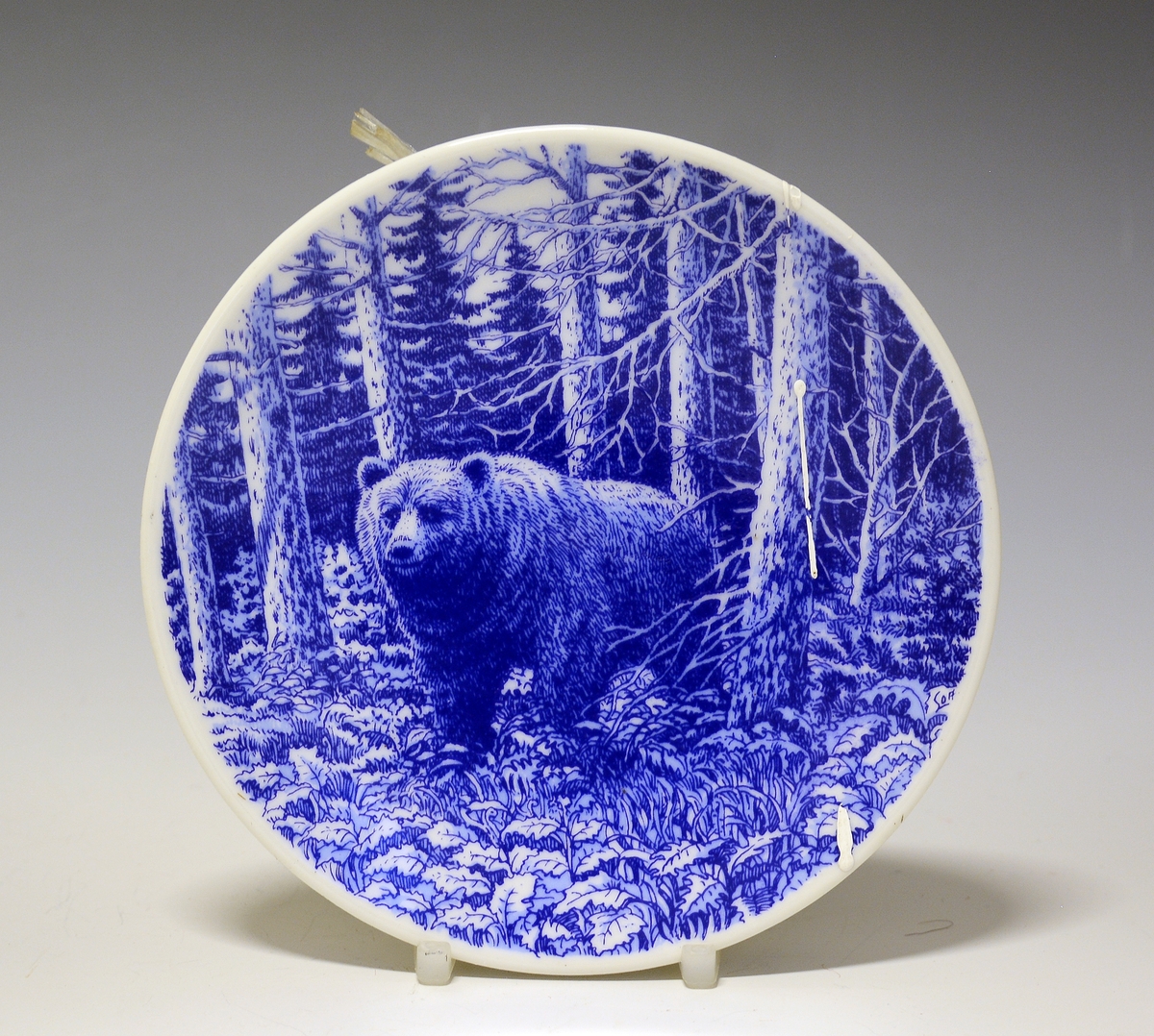 Platte i porselen med blå trykkdekor av bjørn i høstskog. I forgrunnen blader og gress, i mellomgrunnen nakne trær, og i bakgrunnen granskog.