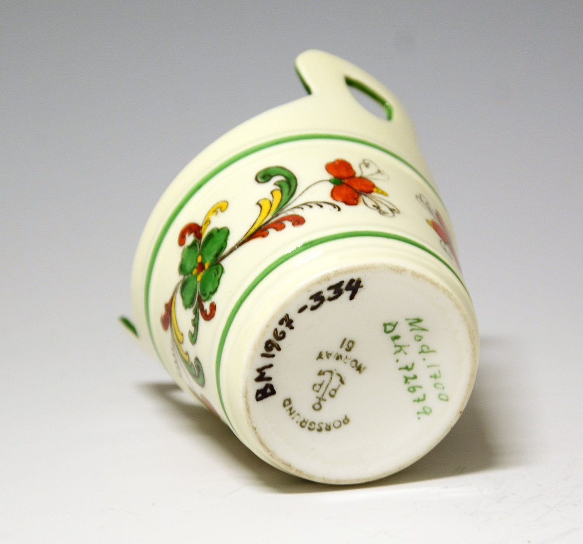 Liten kopp av porselen, formet som en smørstamp. Hvit glasur, også inni. På sidene rosemalt felt i sterke farger.
Modell: 1700
Dekor: 72679