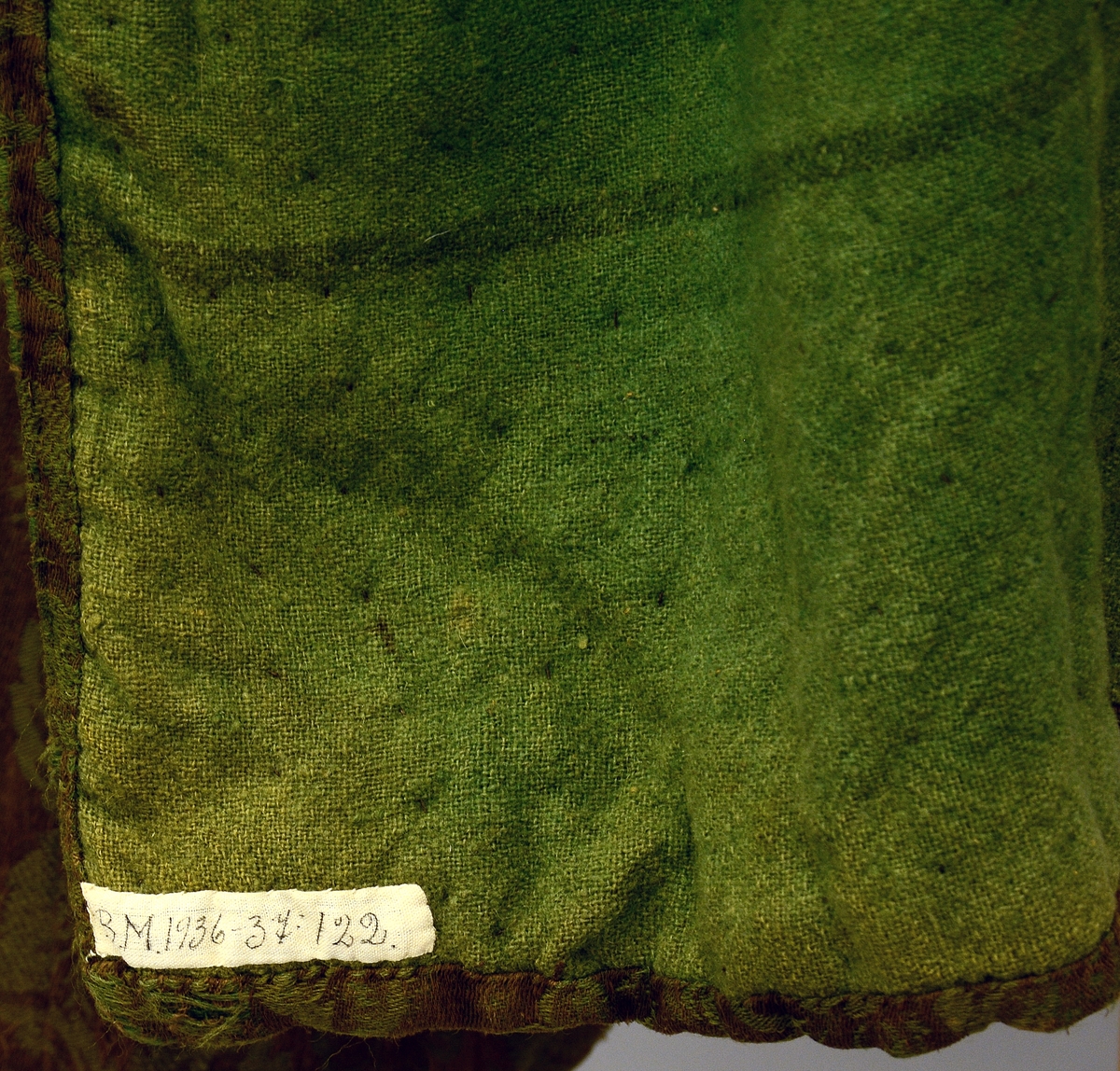 Cape. "Kjerringkaape" av graabrunt bomullsdamask med grøne blomar. Vattera og inni fora med grønt vadmaal. Slengkaape rykt yvi herdane; utan ermar. Fra protokoll.