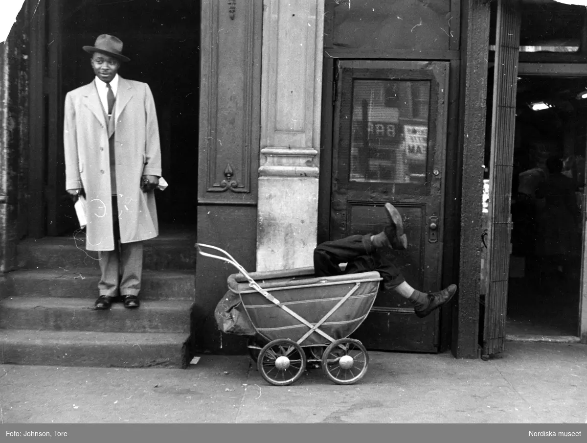 En man i ljus kappa och hatt bredvid en barnvagn där en utslagen man ligger. Lenox Avenue, Harlem, New York, USA.