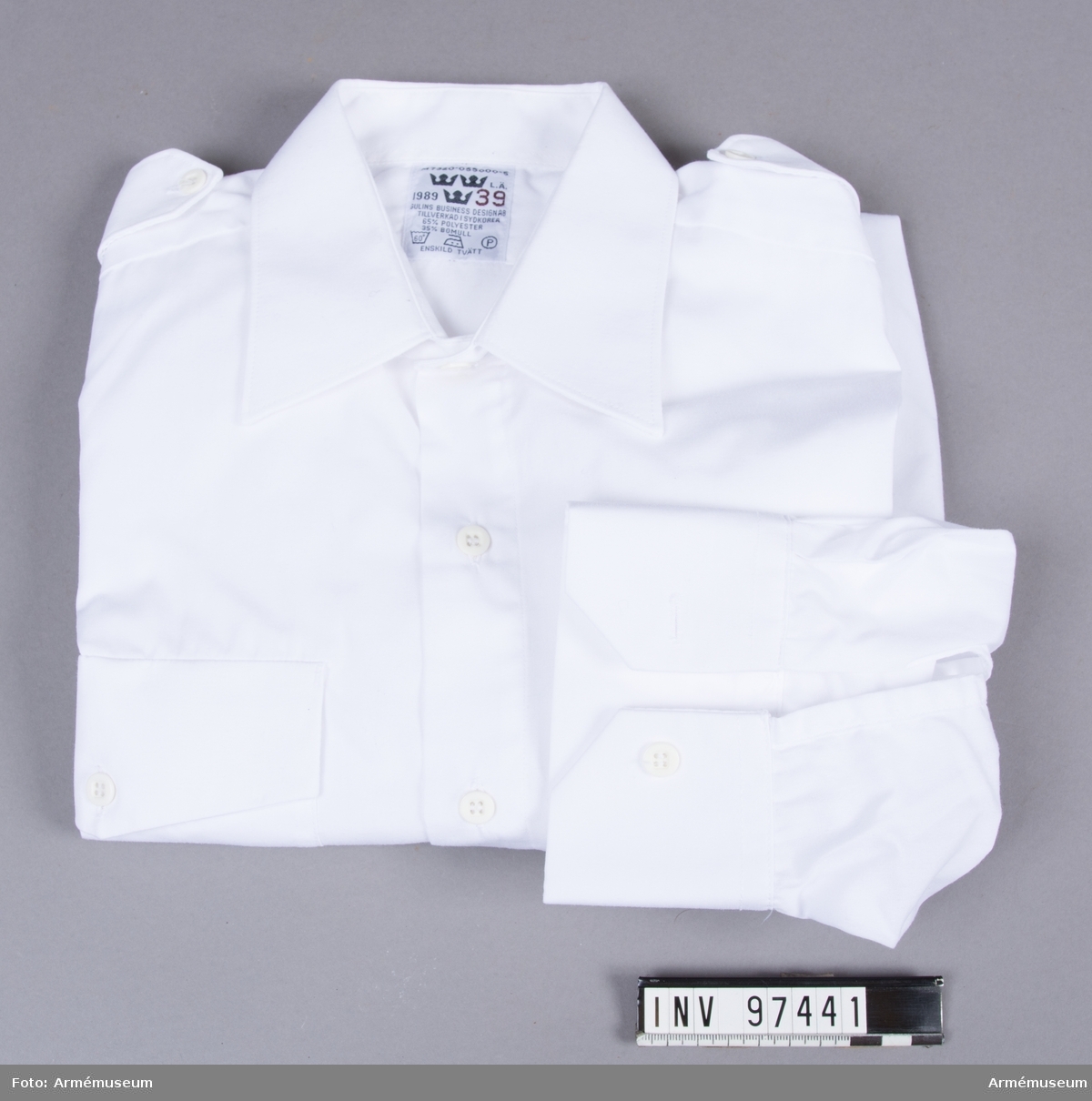 Vit skjorta i polyester och bomull, tillverkad i Sydkorea 1989. Med axelklaffar där axelhylsor kan fästas.
