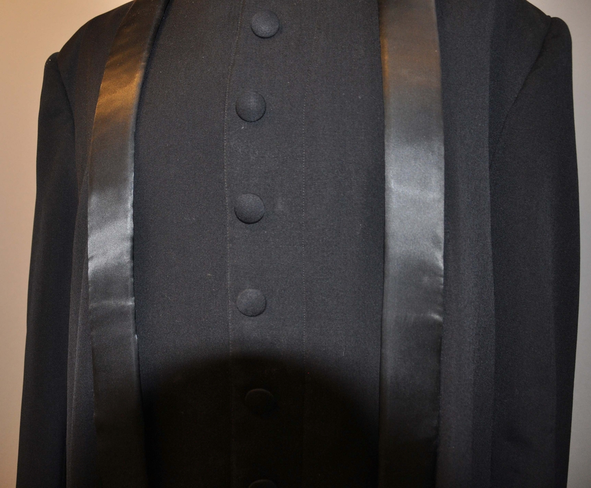 Svart prestekjole med hvite ermer. Har én knapperader på framsiden, samt et silkebånd som går opp langs sidene av knapperaden og rundt kragen.
Den øverste knappen ser ut til å være erstattet. Stoffet er i god stand.