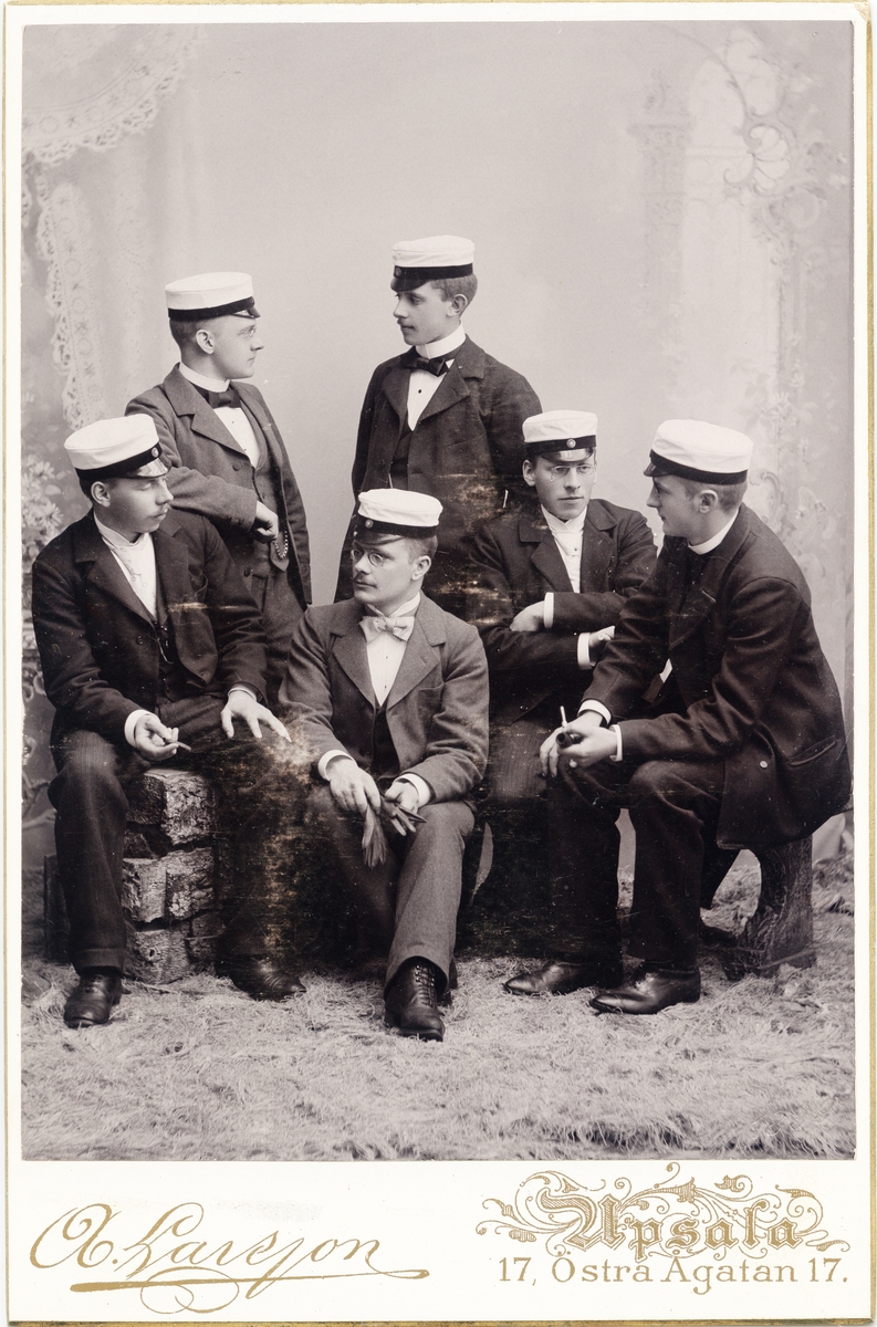Kabinettsfotografi - sex män iklädda kostym och studentmössa, fyra sitter och två står, några håller rökdon i händerna, Uppsala 1899
