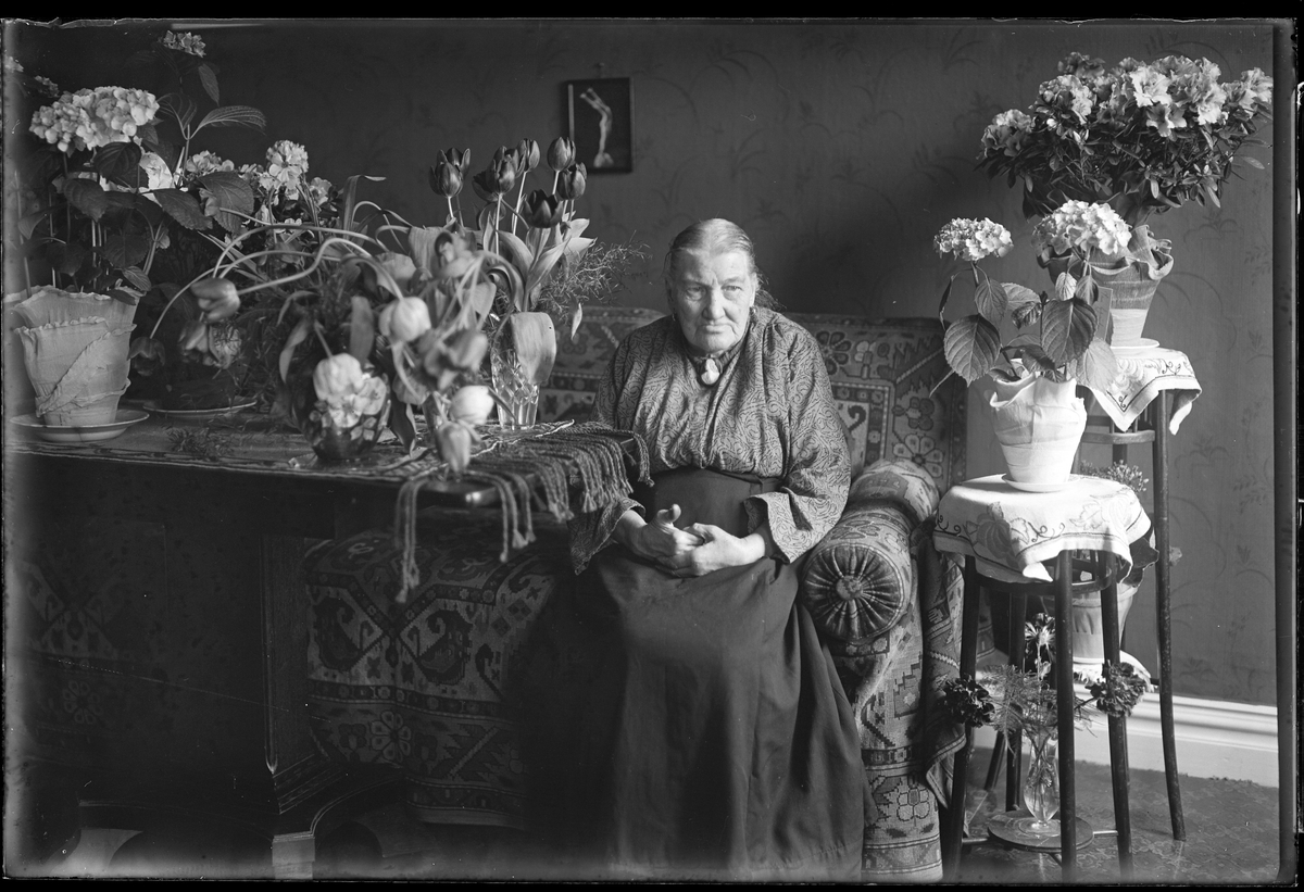 En äldre kvinna, tolkad som Elna Olsdotter, sitter i en soffa omgiven av blommor. Bilden är tagen i samband med hennes 80årsdag.