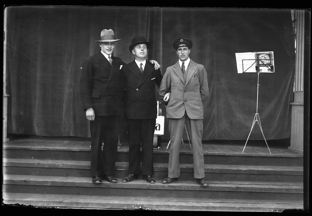 Tre män står på trappan till en scen och håller om varandra. I fotografens anteckningar står det "Gammalmusiken i Parken".