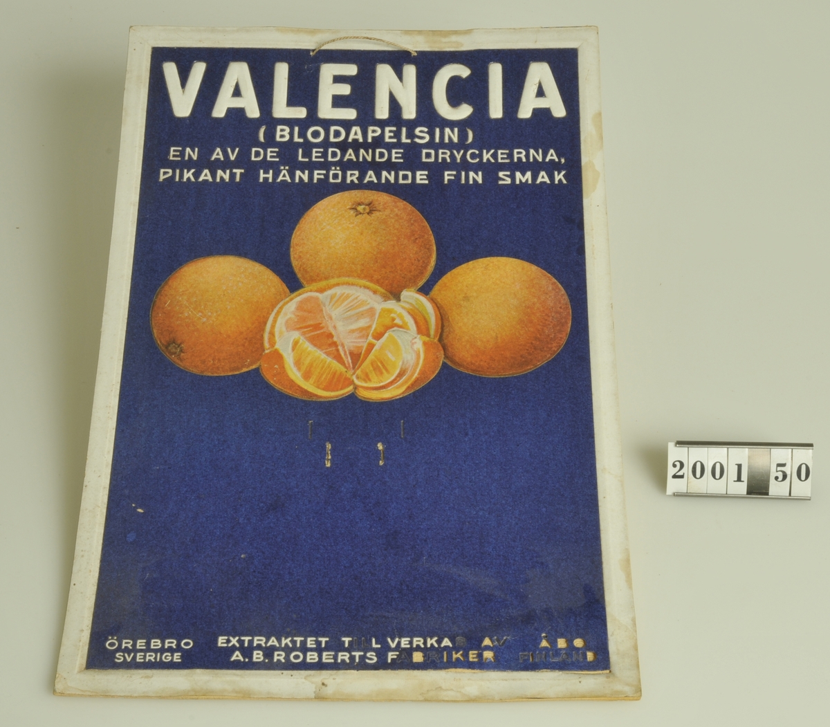 Reklam för Valencia apelsin extrakt. Har märken på mitten som kan betyda att en almanacka varit fäst vid skylten. 
Flera av bokstäverna i den nedre delen har fallit av.


