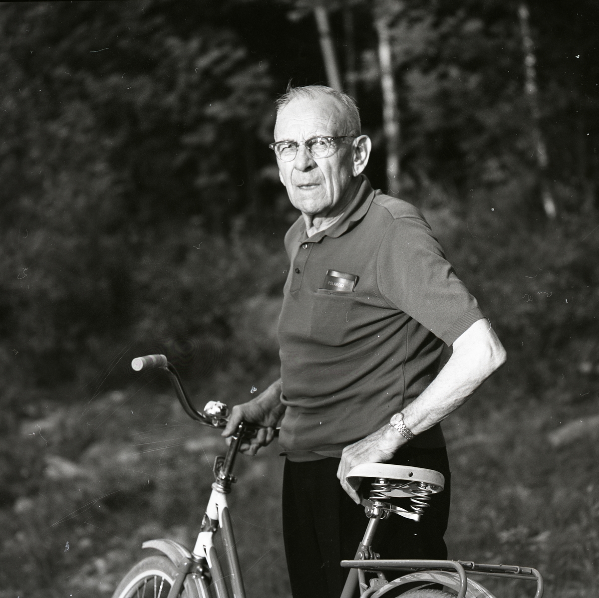 En äldre man i glasögon står med en cykel. I bröstfickan har han sitt glasögonfodral. En mild sol lyser på honom.
