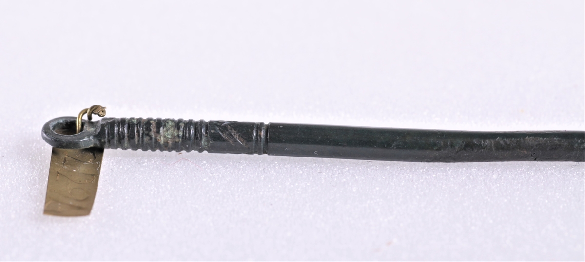 Smykkenål i bronse funnet i en tidligre gravhaug. En lang, spiss nål med et rundt øye. Nålen er dekket av sort emalje som kalles niello.