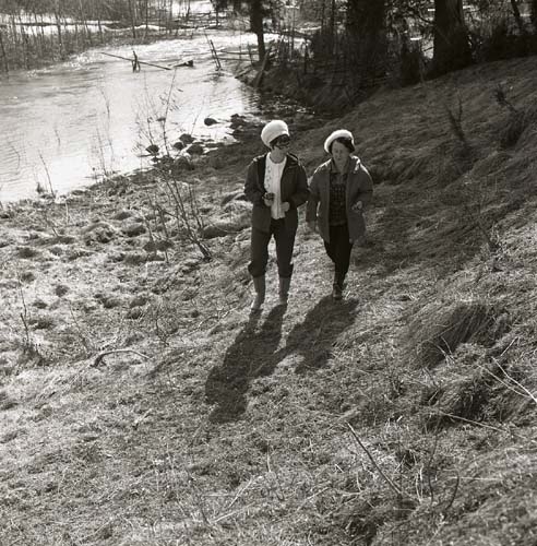 Två kvinnor i ljusa mössor promenerar längs en slänt vid ett vattendrag, 1970.