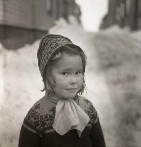 En flicka iförd stickad mössa och tröja står i ett snöigt kvarter, 1953.