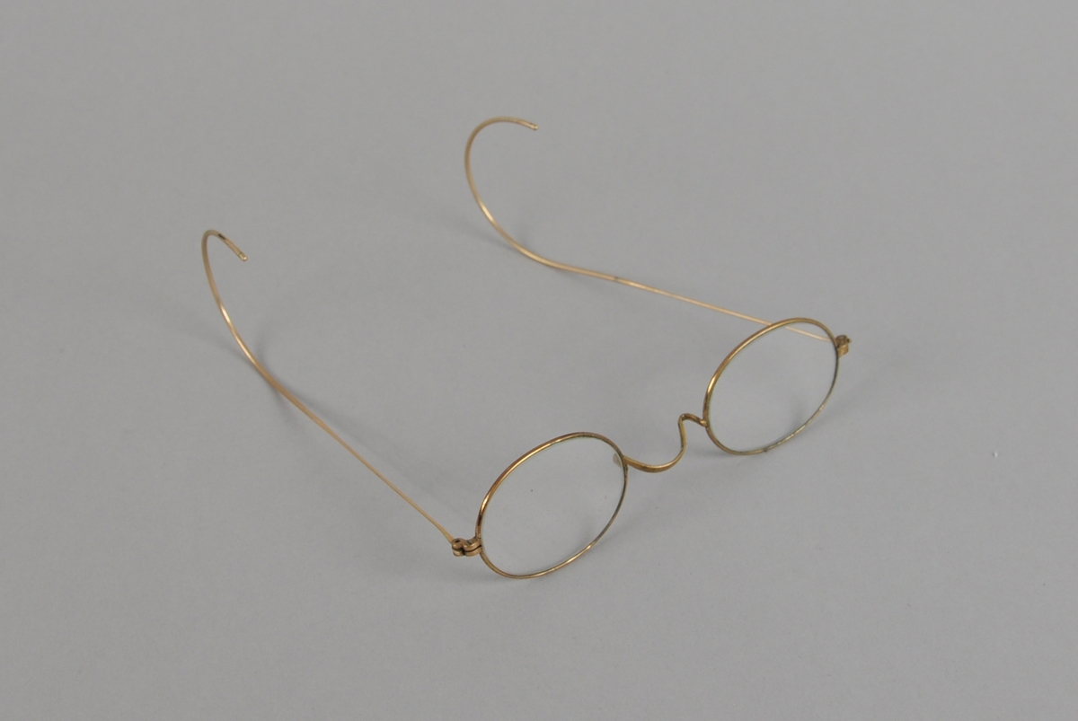 Ovale briller med gullfarget metallinnfatning og buede brillestenger.