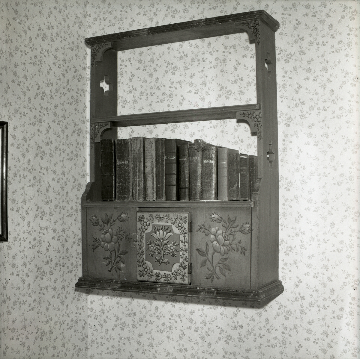 En bokhylla fylld med biblar mot en blommig tapet, 1990.