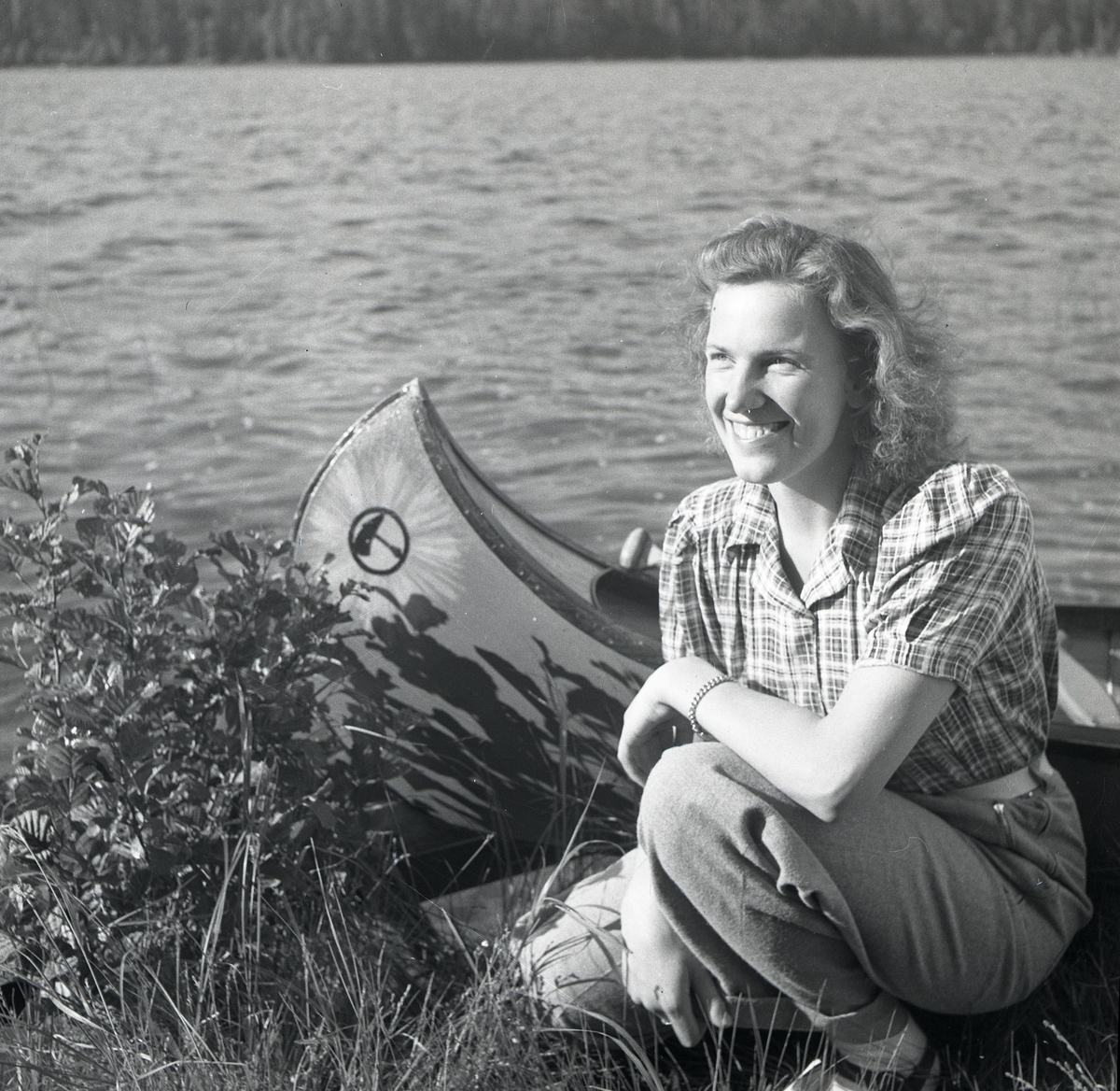 En leende kvinna sitter på huk i gräset vid en sjö bredvid en canadakanot, Skidtjärn 23 juni 1947.