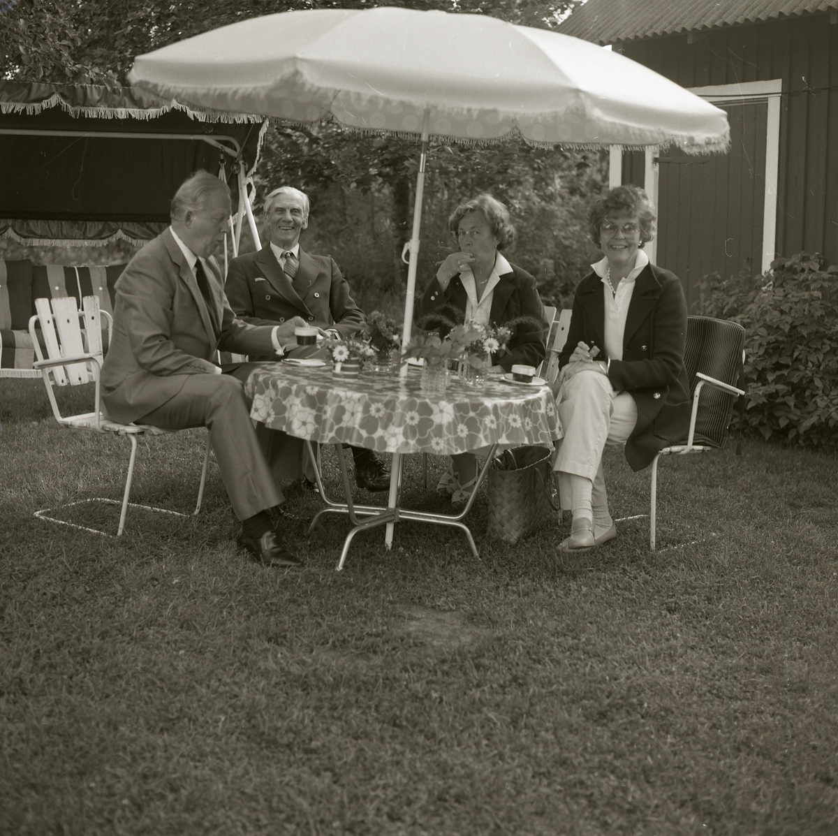Bo Setterlind (t h i bild) hos kyrkoherde Perssons, Långvind 22 juli 1984. Två män och två kvinnor sitter i trädgården under ett parasoll och dricker kaffe. Adéle sitter som tvåa från höger i bild.