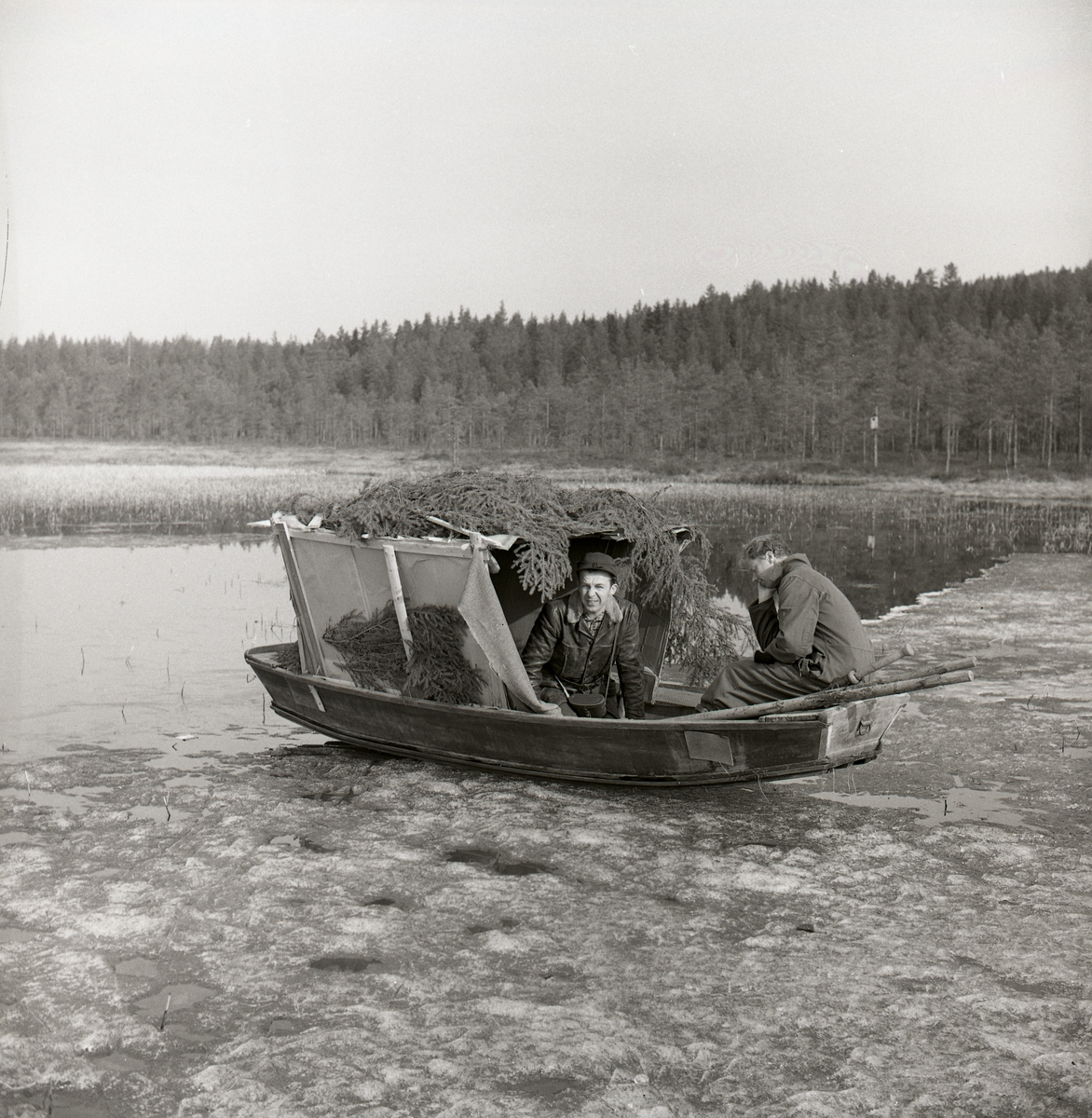 Två män befinner sig vid och i en båt med rumliknande påbyggnad, Stråsjön i april 1964. Båten ligger på en iskant med fören i vattnet.