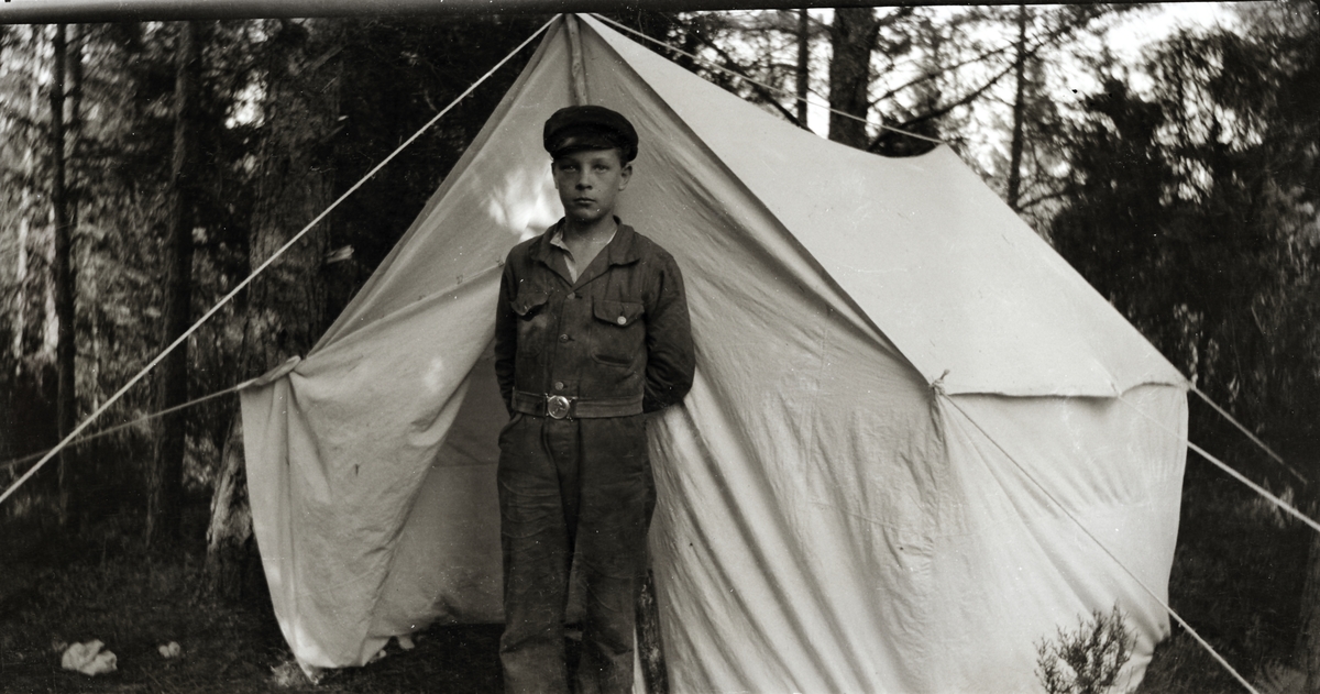 En pojke i overall och vegamössa står med armarna bakom ryggen framför ett tält i skogen.