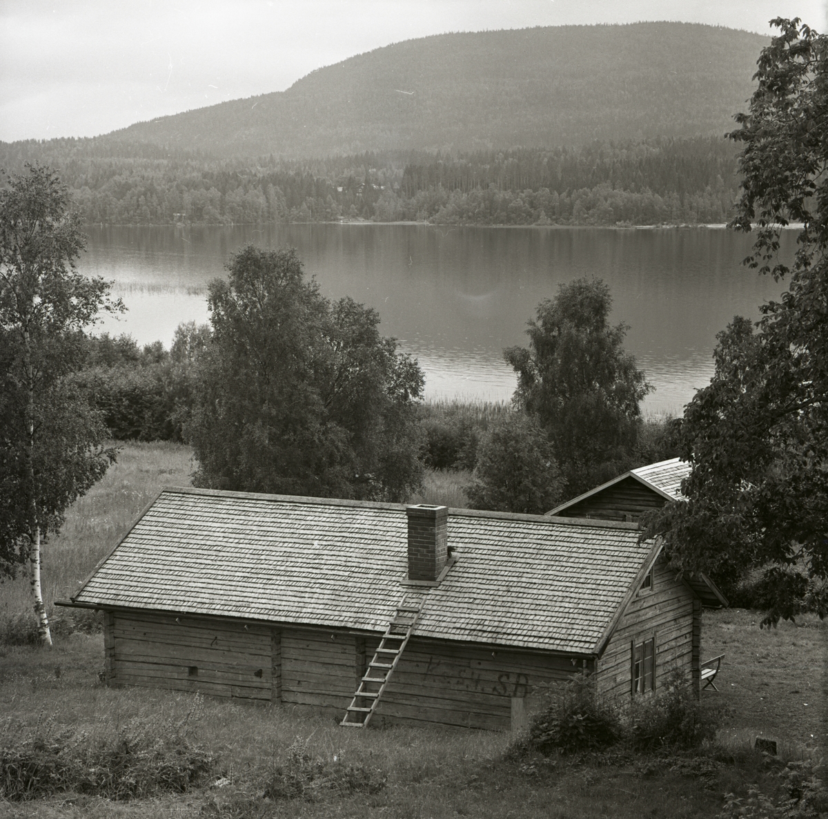 En utsikt över en fäbodstuga med en sjö och ett berg i bakgrunden, 1 augusti 1965 i Älvåsen.