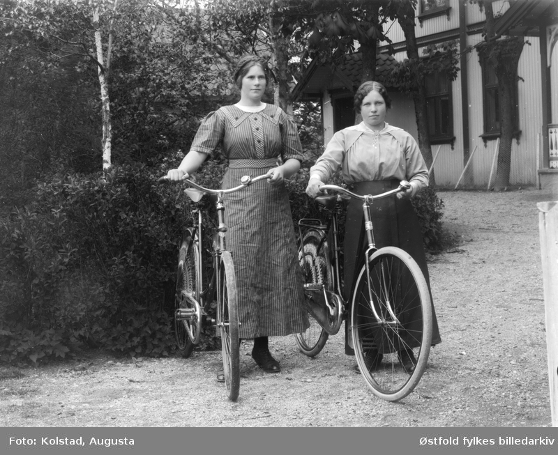 To unge kvinner med sykkel  på tunet utenfor våningshus i Askim ca. 1910 - 1920. Begge kvinnene er ukjente. ØFB.AKO.00022. 