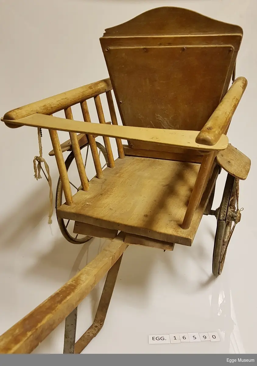 Liten barnestol av tre. Stolen har to hjul av jern og gummi istedet for stolbein. Det er festet en lang trestokk til stolen så den kan dras. Under stanga er det en støtte av jern som stolen hviler på når den ikke er i bruk. Brett mellom armlenene i front.