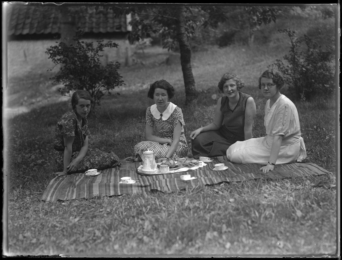 Karin Helgesson, i mönstrad klänning längst ut på kanten, med vänner sitter på en picknickfilt. På filten är kaffekanna, koppar och kakor uppdukade. I fotografens anteckningar står det "Karin Helgesson, Halö, med sällskap". Tolkat som Karin Sofia Helgesson.