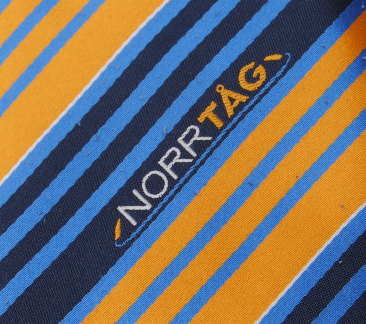 Diagonalmönstrad slips i två olika toner av blått samt gul- och vit färg med broderad text. Slipsen är i två delar och fästs ihop i nacken med hjälp av ett kardborreband. Slipsen är fodrat med en svart textil.(:1) Den smalare delen är försedd med en svart lapp med bland annat tvättråd.(:2)