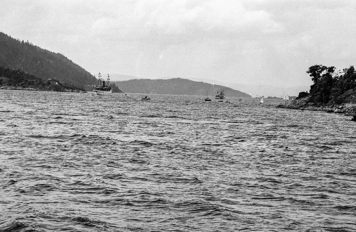 Drøbaksfjorden, sundet ved Oscarsborg. Tre menn på brygga ser på båtene med kikkert. Sandtaket og småhus på Hurum.
Fotograf: ØB Rønning
