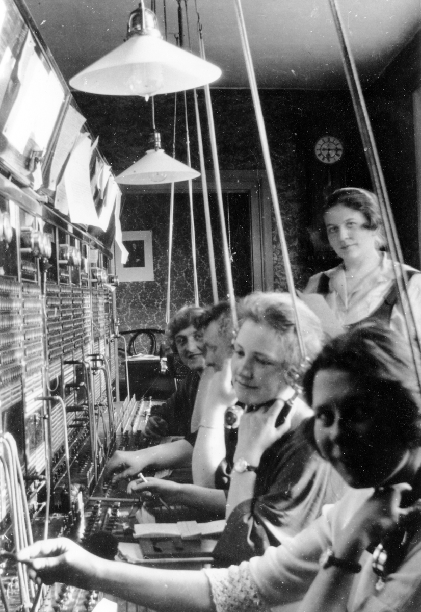Bilden visar fyra kvinnor som sitter och arbetar framför en telegraf och bakom dom en kvinna som står upp. Bilden är tagen på Telegrafstationen på Nygatan 18, kv. Hjorten 19.