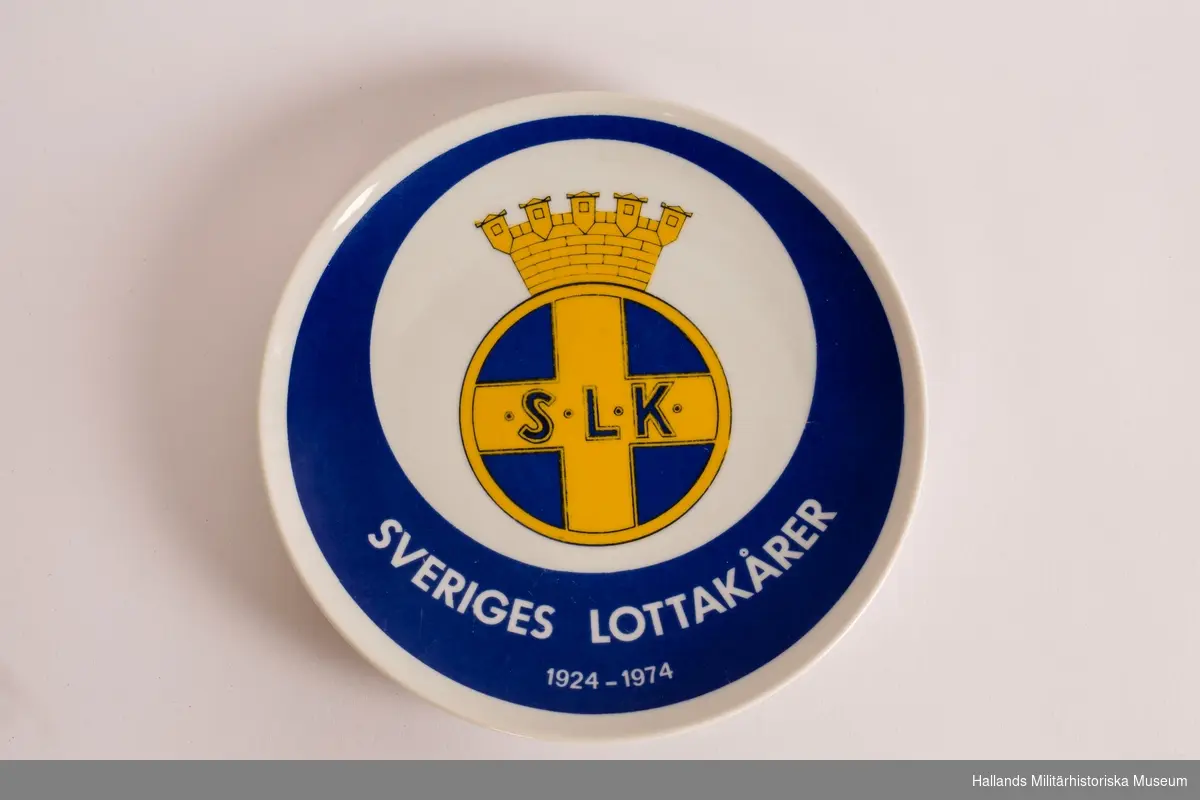 Jubileumstallrik. Tillverkad i porslin. Vit med blå botten. På framsidan Svenska Lottakårens (SLK) emblem samt därunder texten: "SVERIGES LOTTAKÅRER 1924-1974".