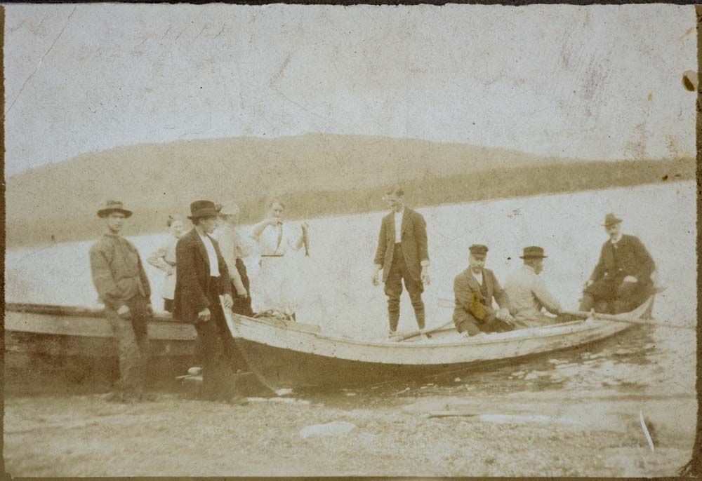 Elvebåter, ukjent sted med ukjente personer både i båtene og stående på land. Kvinne holder opp et par fisk.
