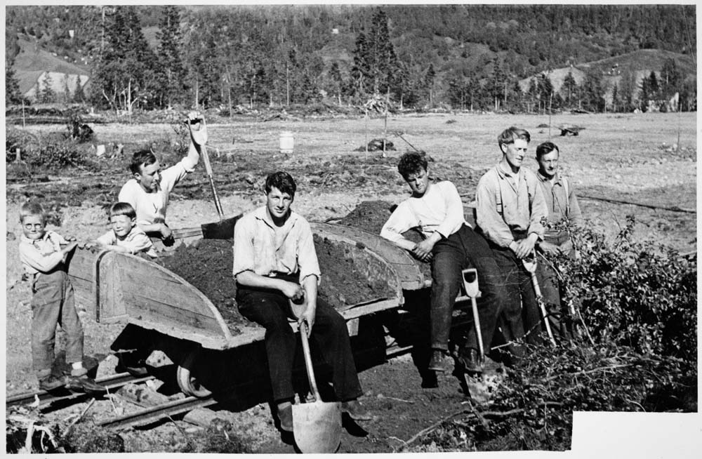 Fem menn med spader graver jord oppi vogn/vagge. To barn.