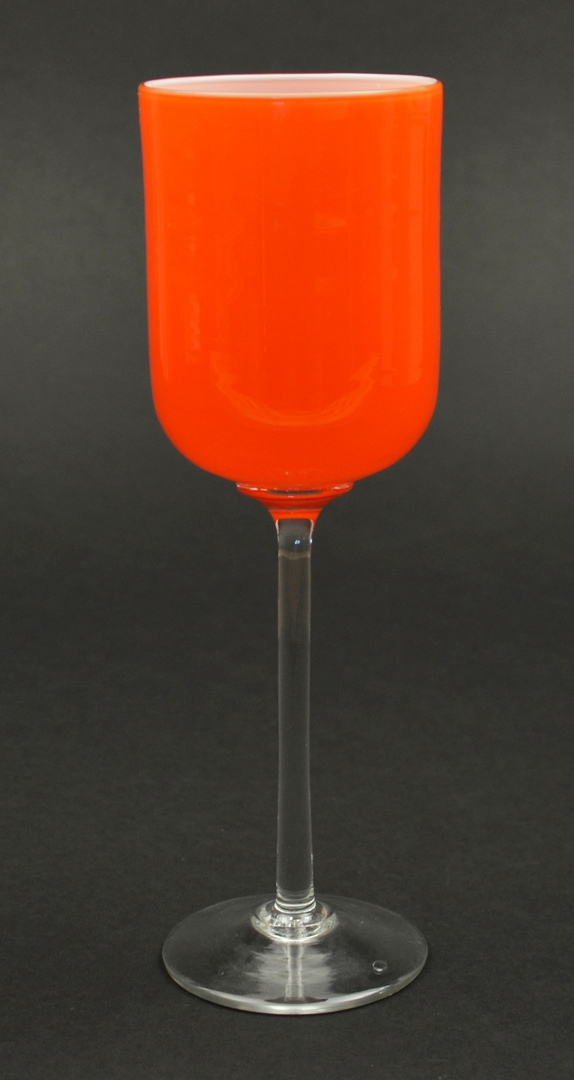 Pokal eller vase på stett.  Handblåse glas. Låg fot og nesten rett stett av fargelaust glas. Sylindrisk klokke av orange glas utanpå og opalint (kvitt) glas inni.