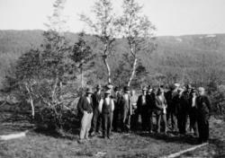 Foto av en gruppe menn med skog og mark i bakgrunnen. Et bil