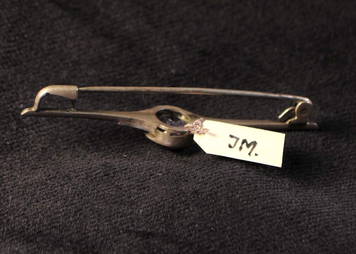 En brosch av silver (?) i form av en nål med infattad lila glassten (ska efterlikna ametist), med markasiter vid sidorna. Slitna stämplar.