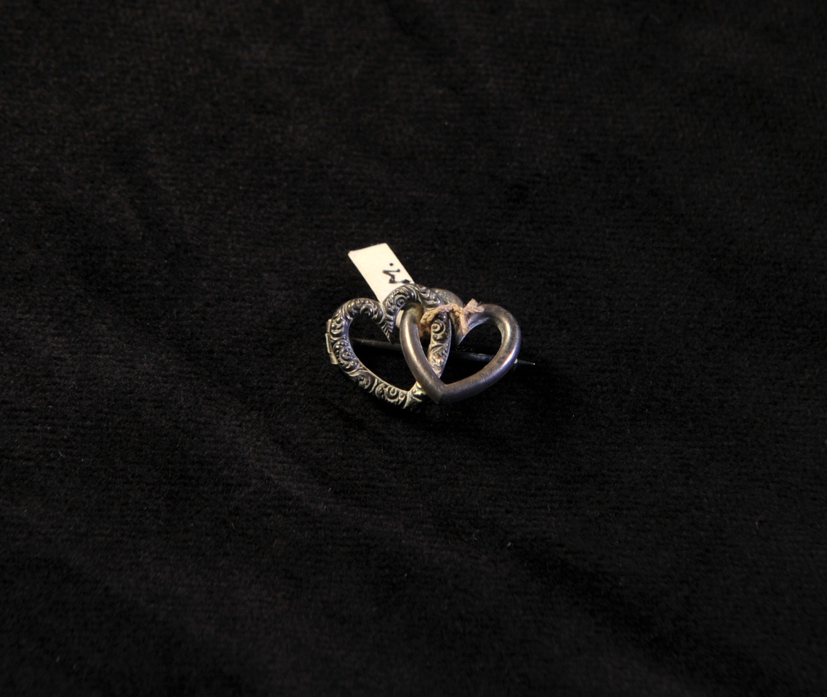 En brosch av silver i form av två st. inflätade hjärtan, ett slätt och ett med akantusdekor. Märkt bakpå: "Sterling