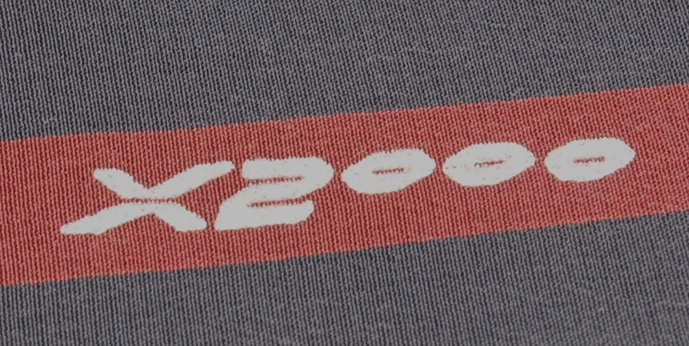 Kvadratisk sidenscarf med rött mönster på svart botten. Mönstret består av röda åttkantiga ovaler, inuti ovalerna finns en rektangulär svart yta. Runt mönstret av ovaler löper en röd kvadratisk ram, och inuti ramen en vit smalare linje. På ett ställe avbryts den smalare vita linjen av texten: "X2000", som också är vit. Utanför ramen fortsätter den svarta bottnen. 

På baksidan av scarfen finns en svart textillapp med texten: "ALL SILK", i vitt. På baksidan av textillappen finns tvättråd, samt texten "MADE IN ITALY". Ovanför denna text står "CONFEZIONATO A MANO", vilket på italienska betyder ungefär: Packad för hand.