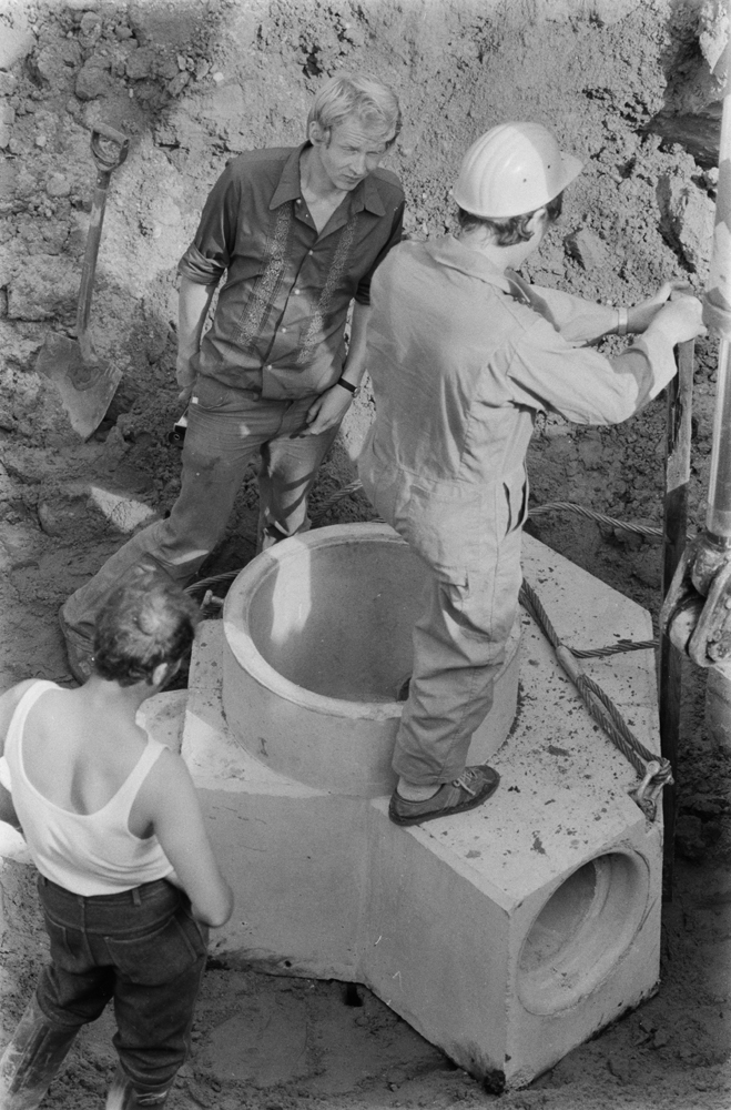 Legging av ny kloakk i Sandnessjøen. Gravemaskin i arbeid. Forgreining i betong blir lagt ned. Flere mann i arbeid.