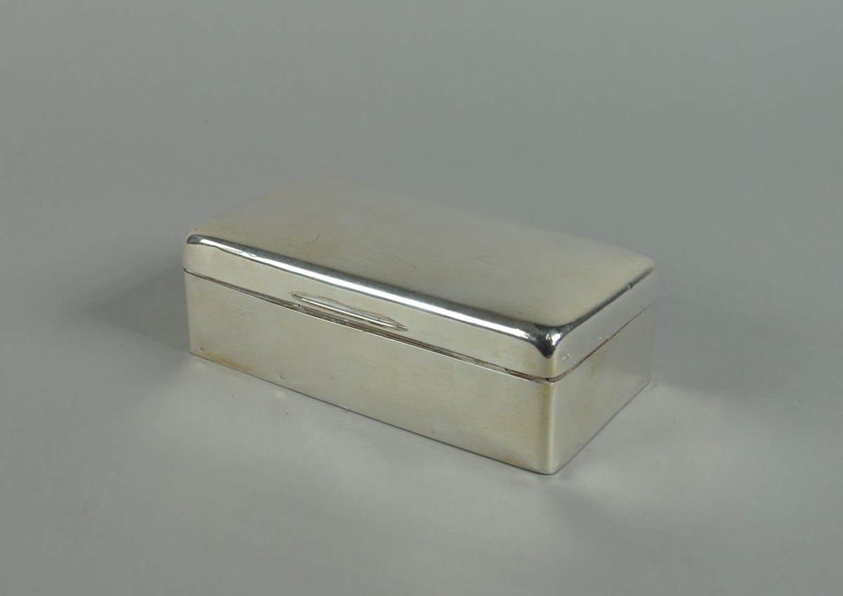 Rektangulært etui av sølv. Etuiet har avrundede hjørner og hengslet lokk. Innsiden av etuiet er innlagt med tre og har to seksjoner.