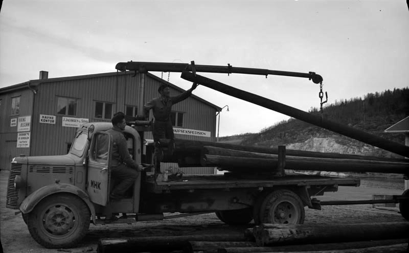 Folk fra Helgeland Kraftlag laster stolper på Jernbanekaia på en lastebil (Bedford O-serie, prod. 1946-53).