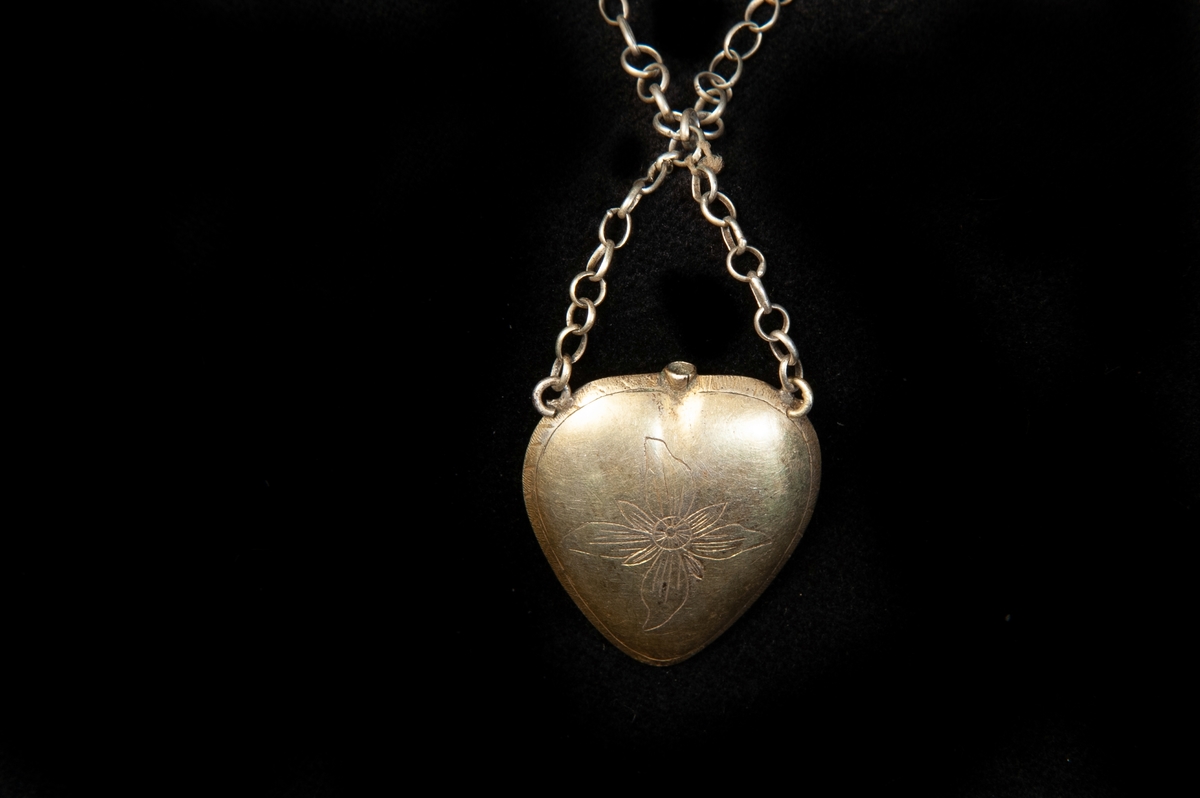 Enkel halskedja av metall med hänge i form av  ett ihåligt hjärta av silver. Graverad dekor i form av en blomma framtill. Baktill en zick-zackbård. Litet hål upptill. Hjärtat har varit avsett för så kallat luktvatten. Stämplad (något otydligt) bakpå.