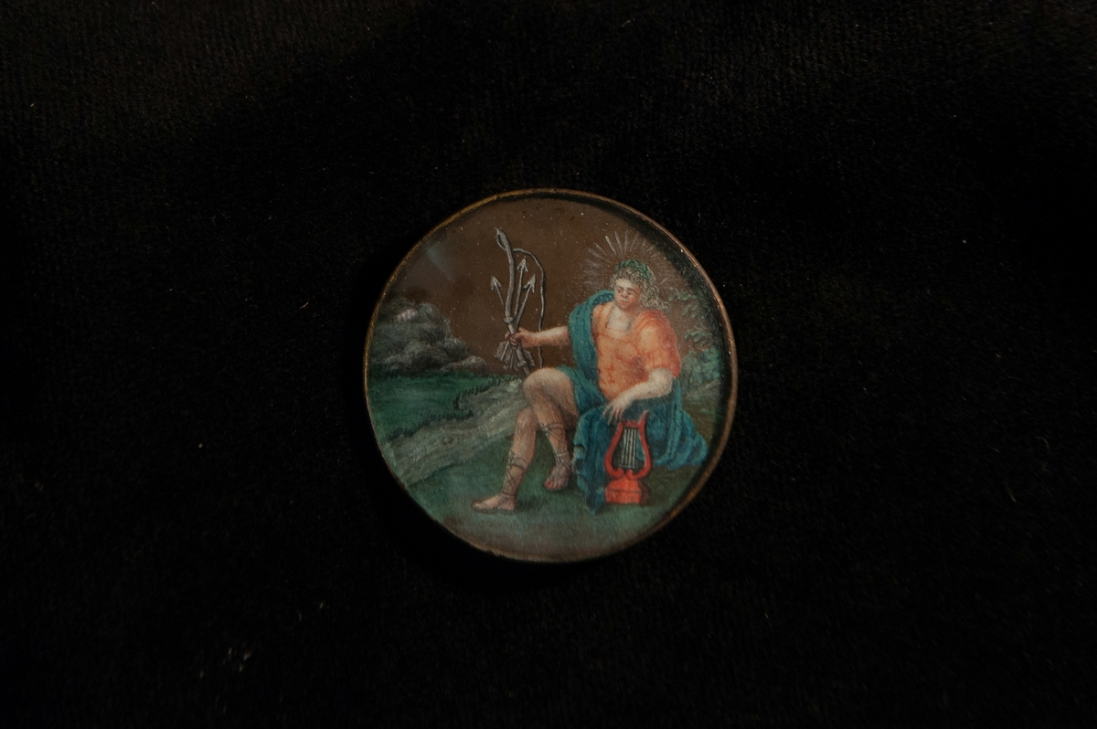 Rund knapp av mässing. Infattad målning bakom glas, föreställande guden Apollon, iförd rosa klädnad med blå mantel m.m., sittande vid en flod. Miniatyr.