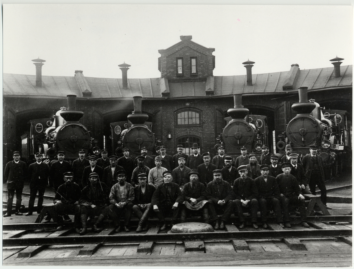 Katrineholms lokstation ca 1895. Personalen poserar framför lokstallet med fyra ånglok i bakgrunden.
Text på bild : I Mitten med helt K. Söderström