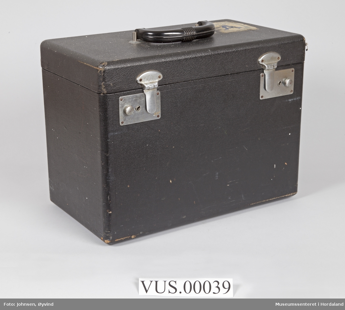 Koffert til Singer Featherweight 221 K Portable Sewing Machine. Eget rom i metall til ekstradeler og trådsneller. Trukket i sort stoff med blomstermotiv i bunnen av kassen. To låser (nøkler mangler), håndtak.
