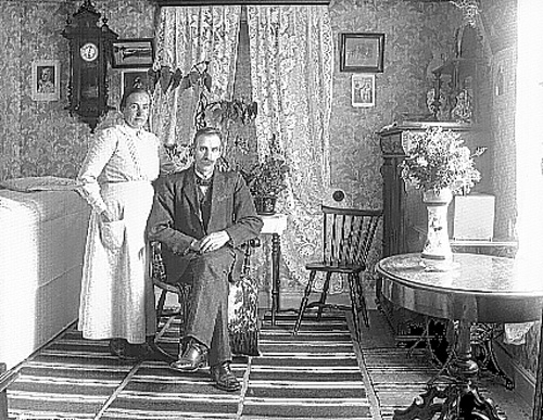 Interiör från snickarmästaren Axel Johansson och hustrun Hanna Ludvigsdotters hem. Hustrun står intill mannen som sitter i en gungstol.